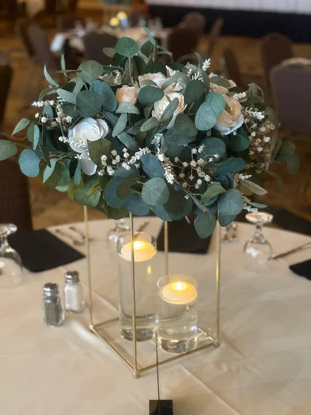 A floral centerpiece with white roses and eucalyptus leaves on a gold stand, accompanied by candles on a white tablecloth.