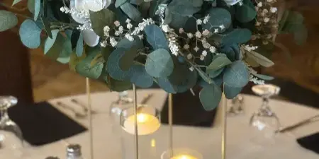 A floral centerpiece with white roses and eucalyptus leaves on a gold stand, accompanied by candles on a white tablecloth.