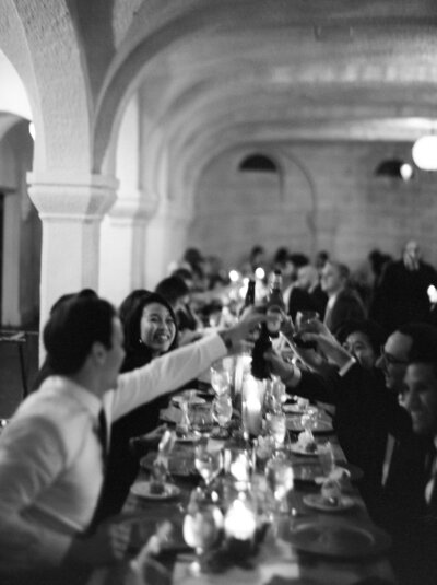 Wedding guests raising glasses in a toast at a long banquet table in an arched venue.
