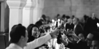 Wedding guests raising glasses in a toast at a long banquet table in an arched venue.
