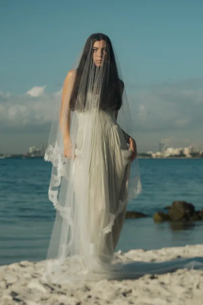 A bride in a white wedding dress and long veil standing on a rocky shoreline with the ocean and a distant city skyline in the background.