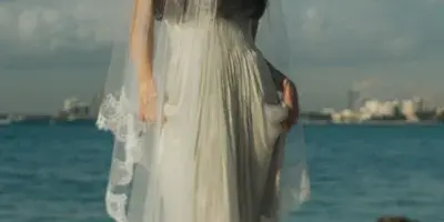 A bride in a white wedding dress and long veil standing on a rocky shoreline with the ocean and a distant city skyline in the background.