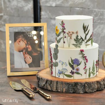 A two-tier white wedding cake decorated with hand-painted wildflowers and greenery, displayed on a wooden slab next to a framed couple photo and cake server.