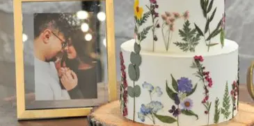 A two-tier white wedding cake decorated with hand-painted wildflowers and greenery, displayed on a wooden slab next to a framed couple photo and cake server.