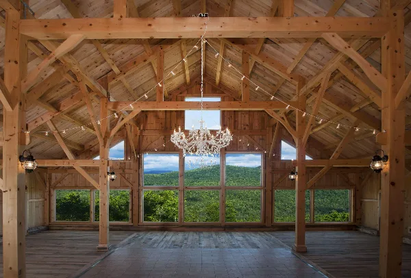 Interior view of a timber frame barn with a chandelier and large windows overlooking a mountain landscape.