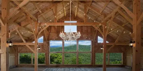Interior view of a timber frame barn with a chandelier and large windows overlooking a mountain landscape.