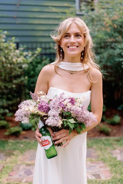 A smiling blonde bride in a white dress holding a bouquet of purple and white flowers and a green bottle of Stella Artois.