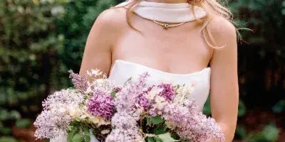 A smiling blonde bride in a white dress holding a bouquet of purple and white flowers and a green bottle of Stella Artois.