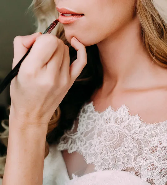 Close-up of a makeup artist applying lip gloss to a bride in a white lace wedding dress.