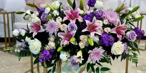 A large, elaborate floral arrangement with pink lilies, purple roses, and white flowers displayed on a gold stand.