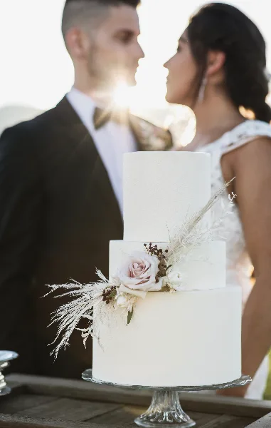 White tiered wedding cake with floral accents and newlyweds behind it