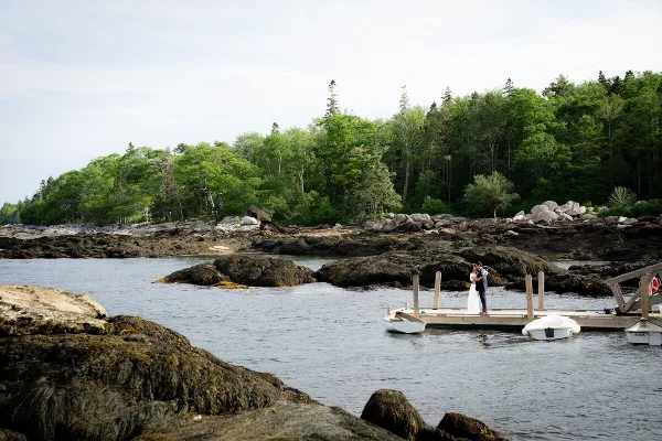 A bride and groom standing on a wooden dock in a coastal landscape with rocky shores and trees.