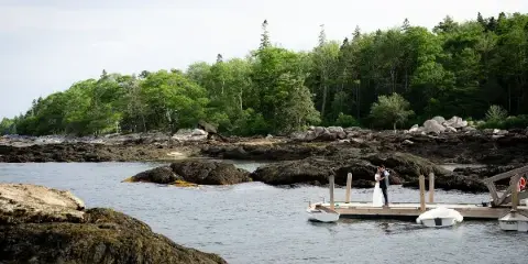 A bride and groom standing on a wooden dock in a coastal landscape with rocky shores and trees.