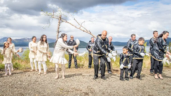 A group of people in traditional Alutiiq and Yup'ik clothing performing a dance outdoors near a body of water in Alaska.