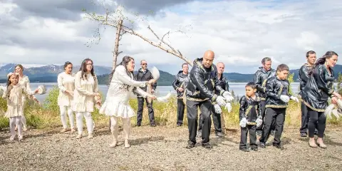 A group of people in traditional Alutiiq and Yup'ik clothing performing a dance outdoors near a body of water in Alaska.