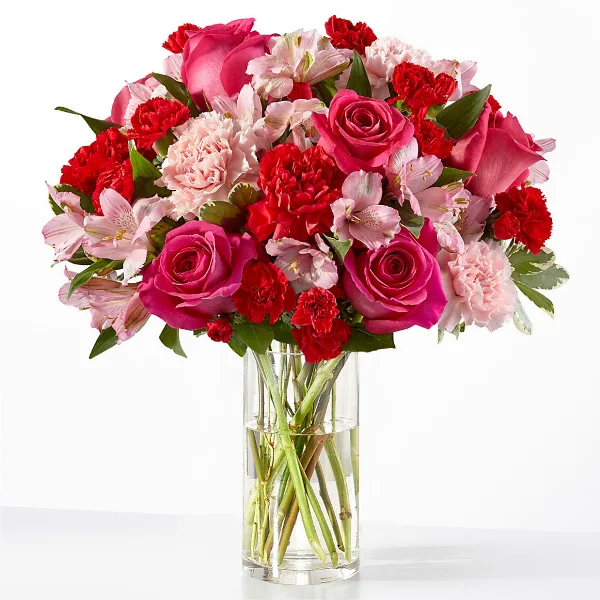 A vibrant floral bouquet featuring pink roses, red carnations, and pink alstroemeria arranged in a clear glass cylinder vase.