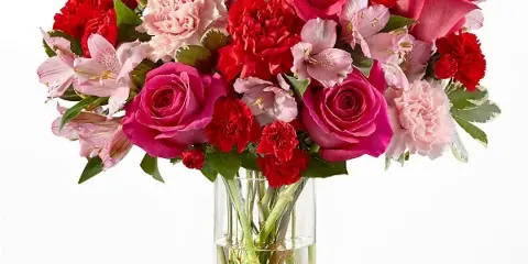 A vibrant floral bouquet featuring pink roses, red carnations, and pink alstroemeria arranged in a clear glass cylinder vase.