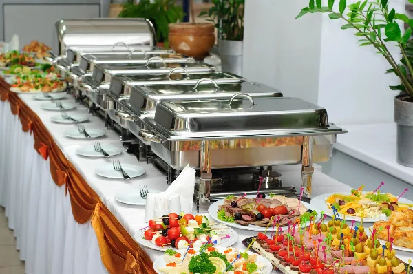 A catering buffet table featuring stainless steel chafing dishes, plates, and various prepared food items.