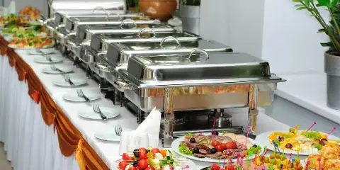 A catering buffet table featuring stainless steel chafing dishes, plates, and various prepared food items.