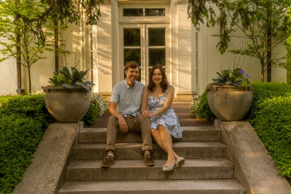 A smiling couple seated on stone steps in front of a white building with large planters and greenery.
