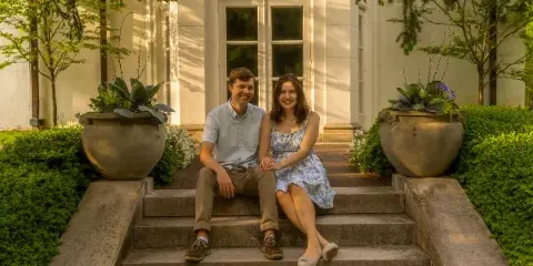A smiling couple seated on stone steps in front of a white building with large planters and greenery.