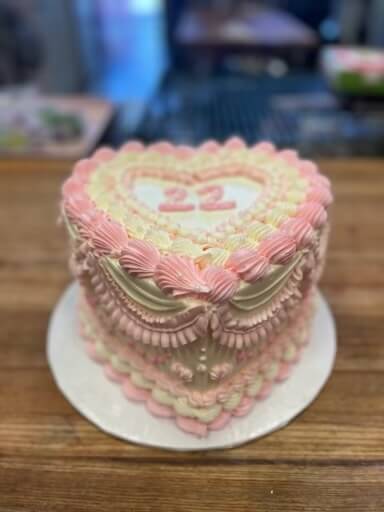 A custom two-tier heart-shaped cake with pink and cream frosting and the number 22 on top, sitting on a white cake stand.