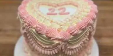 A custom two-tier heart-shaped cake with pink and cream frosting and the number 22 on top, sitting on a white cake stand.