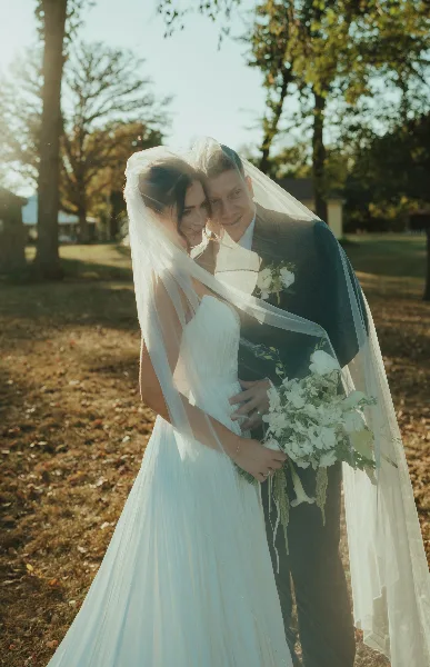 A bride and groom standing together outdoors, the bride holding a white bouquet and wearing a veil, with the groom in a dark suit.