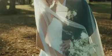 A bride and groom standing together outdoors, the bride holding a white bouquet and wearing a veil, with the groom in a dark suit.