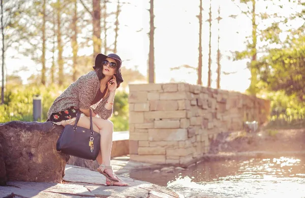 A woman in a hat and cardigan posing with a handbag outdoors near a stone wall and water.