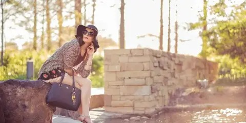 A woman in a hat and cardigan posing with a handbag outdoors near a stone wall and water.
