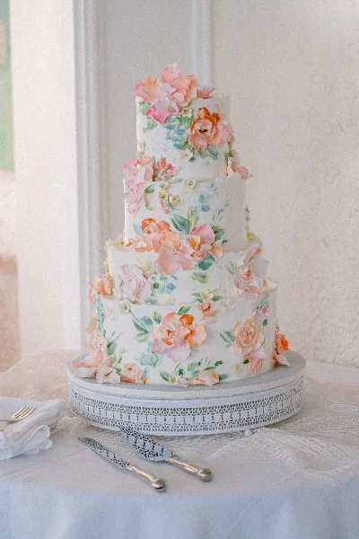 A four-tier white wedding cake decorated with pink and orange sugar flowers and green leaves, displayed on a silver cake stand with serving utensils.