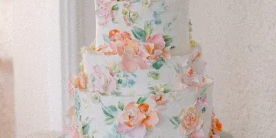 A four-tier white wedding cake decorated with pink and orange sugar flowers and green leaves, displayed on a silver cake stand with serving utensils.