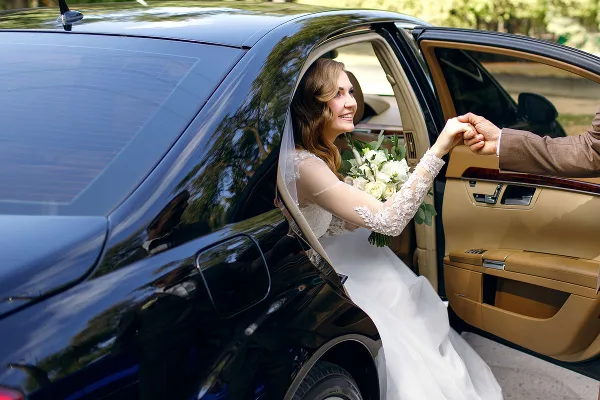 A bride in a white gown and veil is being helped into the back seat of a black luxury SUV by a driver.
