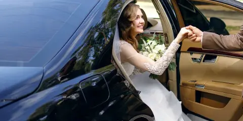 A bride in a white gown and veil is being helped into the back seat of a black luxury SUV by a driver.