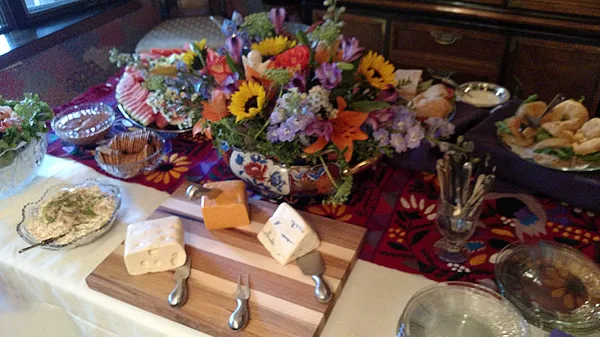 A catering table setting featuring a wooden cheese board, a large floral centerpiece, and assorted food platters.