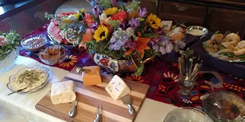A catering table setting featuring a wooden cheese board, a large floral centerpiece, and assorted food platters.