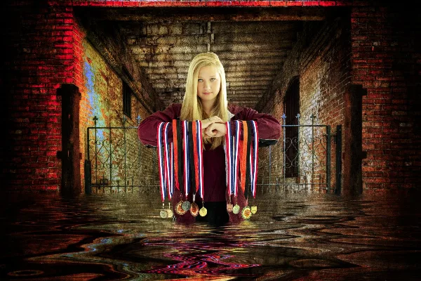 Portrait of a young woman with long blonde hair wearing a maroon top and holding athletic medals, posed in a brick corridor with wet floor reflections.