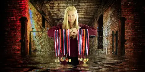 Portrait of a young woman with long blonde hair wearing a maroon top and holding athletic medals, posed in a brick corridor with wet floor reflections.