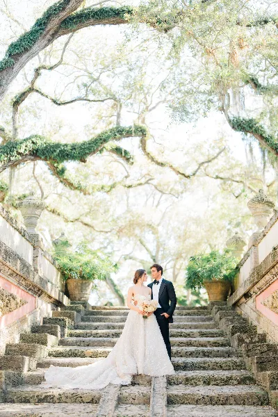Bride in a white gown and groom in a tuxedo standing on stone steps beneath an ivy-covered tree at an estate.