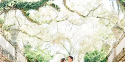 Bride in a white gown and groom in a tuxedo standing on stone steps beneath an ivy-covered tree at an estate.