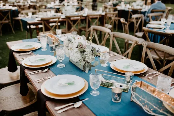 A long wooden table set with blue runners, white plates, and floral centerpieces, surrounded by vintage wooden cross-back chairs.
