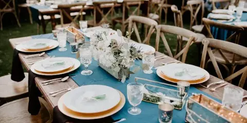 A long wooden table set with blue runners, white plates, and floral centerpieces, surrounded by vintage wooden cross-back chairs.