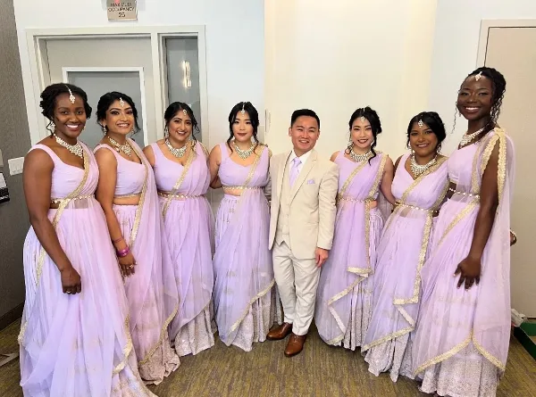 A group of women in lavender South Asian bridal attire with styled hair and makeup posing with a man in a suit.