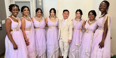 A group of women in lavender South Asian bridal attire with styled hair and makeup posing with a man in a suit.