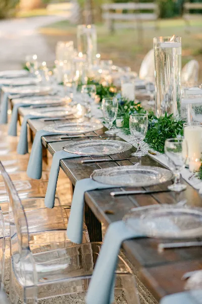 An outdoor wedding reception table set with silver charger plates, glassware, and a green floral runner.