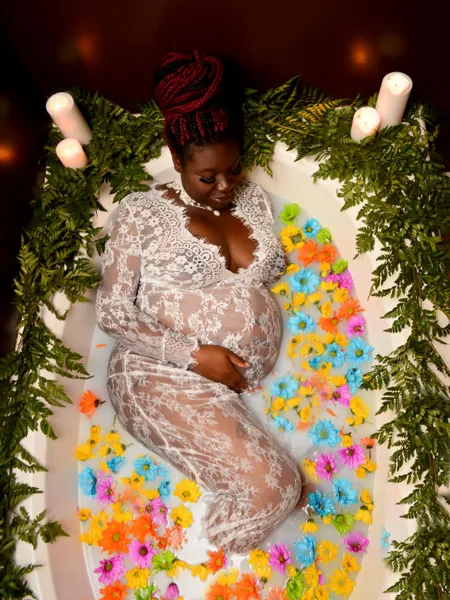 Pregnant woman in a white lace gown lying in a bathtub filled with water and colorful flowers, surrounded by greenery and candles.