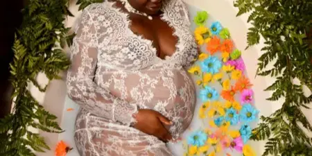 Pregnant woman in a white lace gown lying in a bathtub filled with water and colorful flowers, surrounded by greenery and candles.