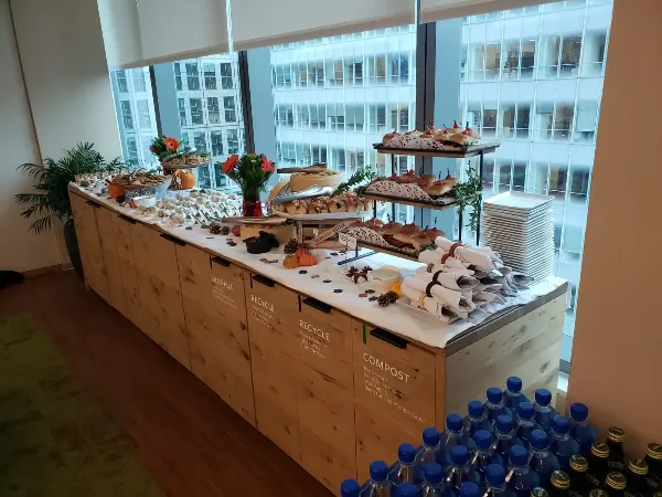 A catering buffet table set up in an office space, featuring various food trays, sandwiches, and water bottles.