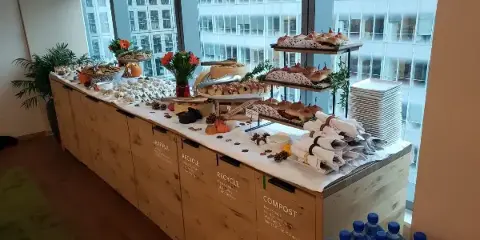 A catering buffet table set up in an office space, featuring various food trays, sandwiches, and water bottles.